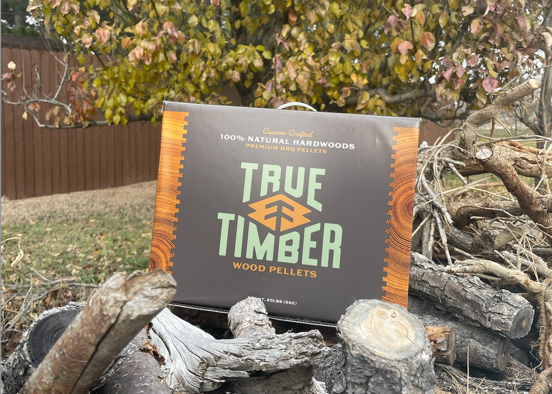 True Timber 100% natural hardwood wood pellets box sitting on a pile of logs outdoors